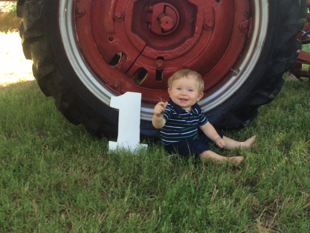 One year old boy sitting in front of a red tractor tire next to a white cardboard cutout of a number one
