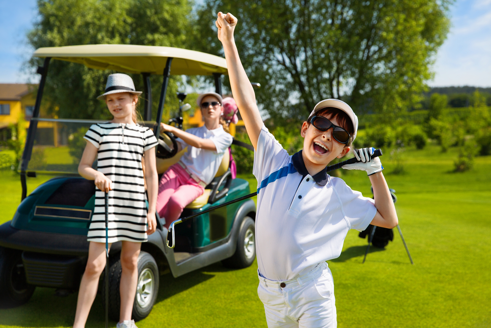 Boy holding golf club cheering with two girls in the background with golf cart