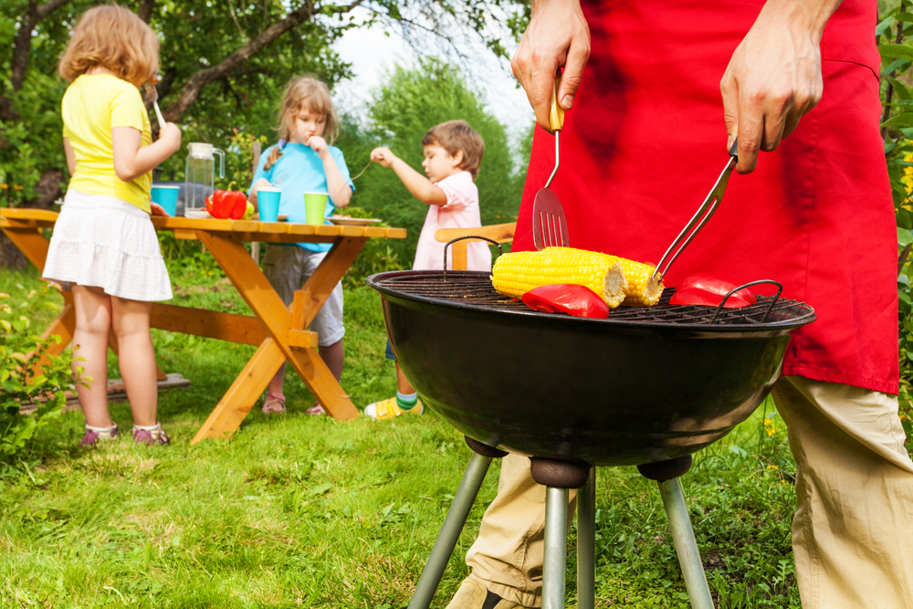 Man in red apron grilling vegetables on grill with kids in background