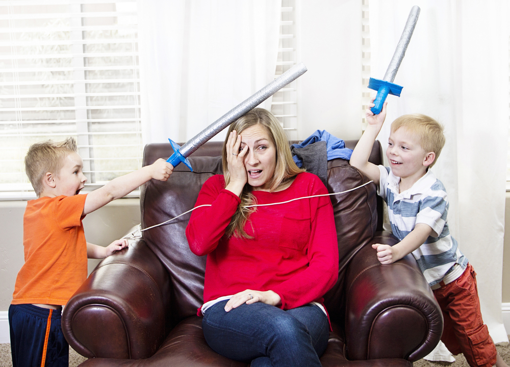 Overwhelmed looking mom sitting in a chair with two young boys on either sided of her pretending to hit her with toy swords