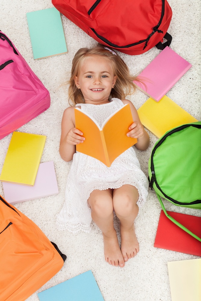 Preschool girl lying on floor looking at book with colorful books and backpacks around her