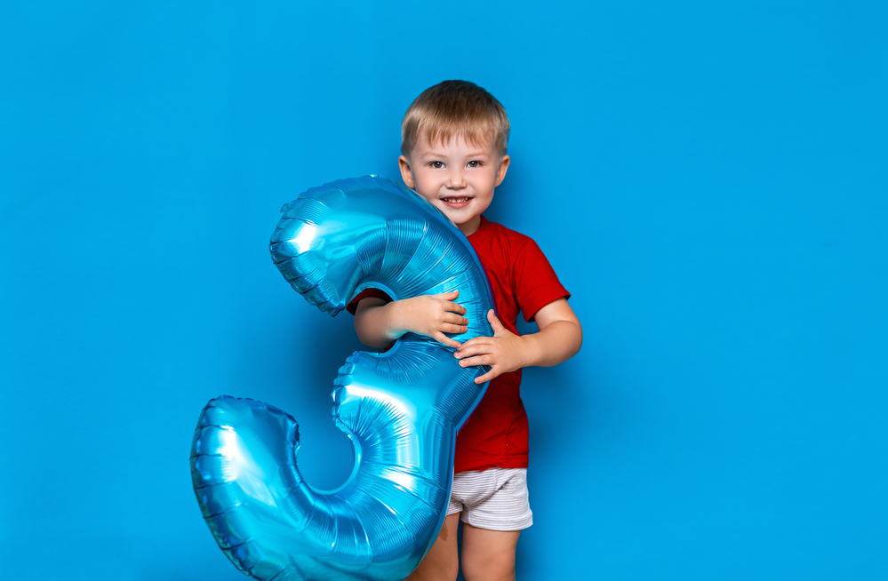 Three year old boy holding a huge blown up foil coated blue balloon shaped like the number three