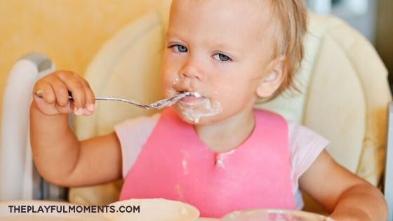 Toddler Girl Sitting in a High Chair Eating With a Spoon