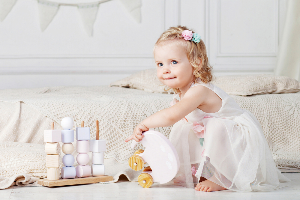 Toddler girl playing with pastel wooden blocks on floor beside a bed with a tan bedspread